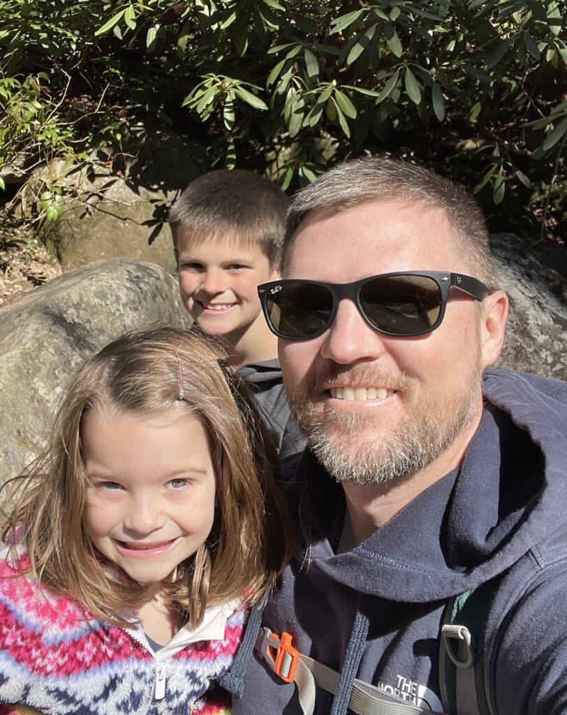 Dad smiles at the camera while hiking with his two children